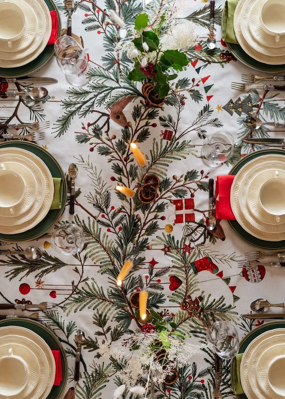 Table de Noël décorée avec une nappe Tessitura Toscana à motifs de sapins et feuillages, couverts et vaisselle blanche.