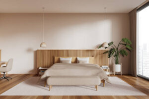 Intérieur de chambre moderne aux tons beige avec parquet en bois, grand lit king size habillé de linge de lit élégant, tables de chevet et plante verte près d’une large fenêtre panoramique.